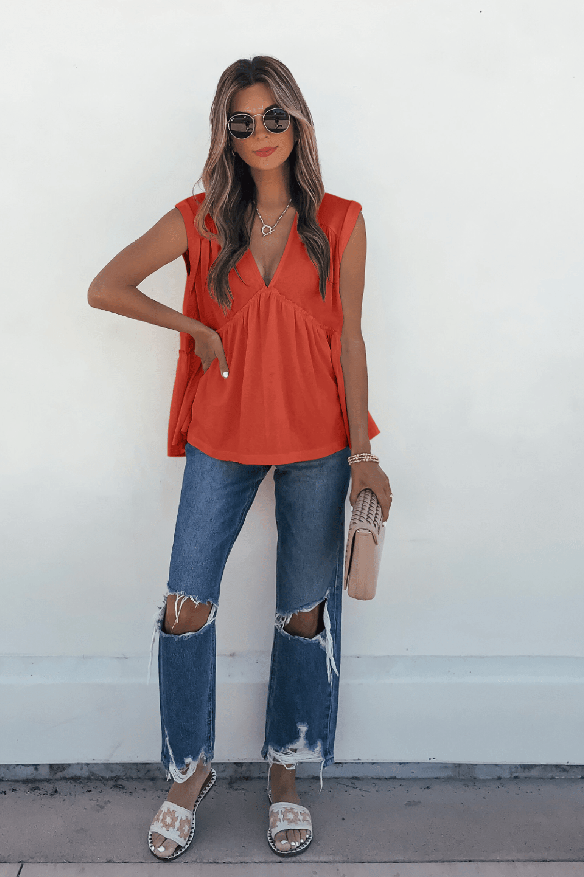 Woman in sunglasses wears Lena's Carefree Afternoon Coral Babydoll Top, ripped jeans, and holds a beige clutch against a white wall.