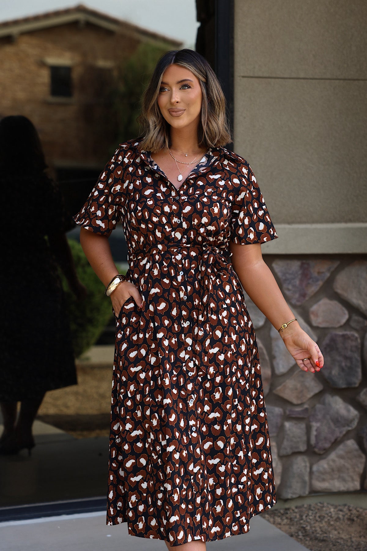 Woman wears the Leopard Print Belted Midi Dress - Brown, smiling outside with one hand in her pocket and the other relaxed by her side.