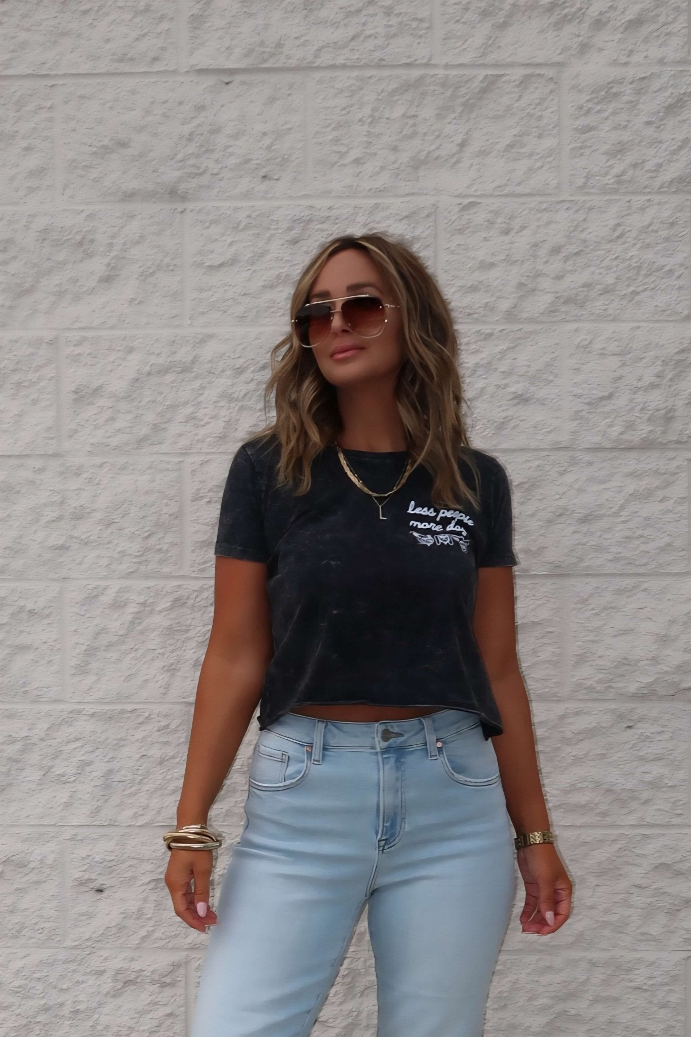Woman in sunglasses, a black Less People More Dogs Graphic Tee, and light blue jeans stands against a textured brick wall.