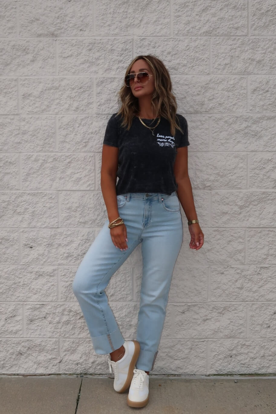 Woman wearing sunglasses, a black Less People More Dogs Graphic Tee, light blue jeans, and white sneakers poses by a brick wall.