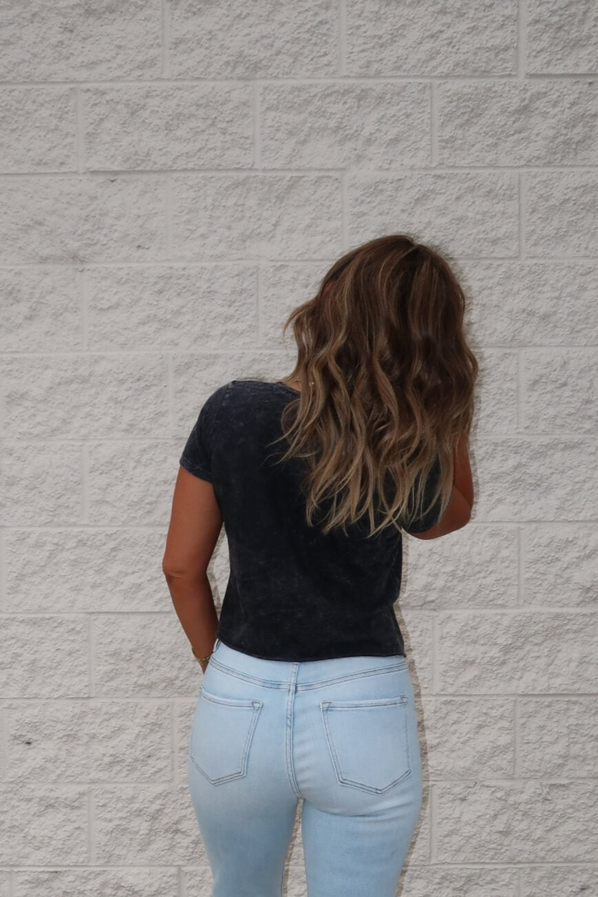 A woman in a black Less People More Dogs Graphic Tee and light blue jeans stands facing a white brick wall.