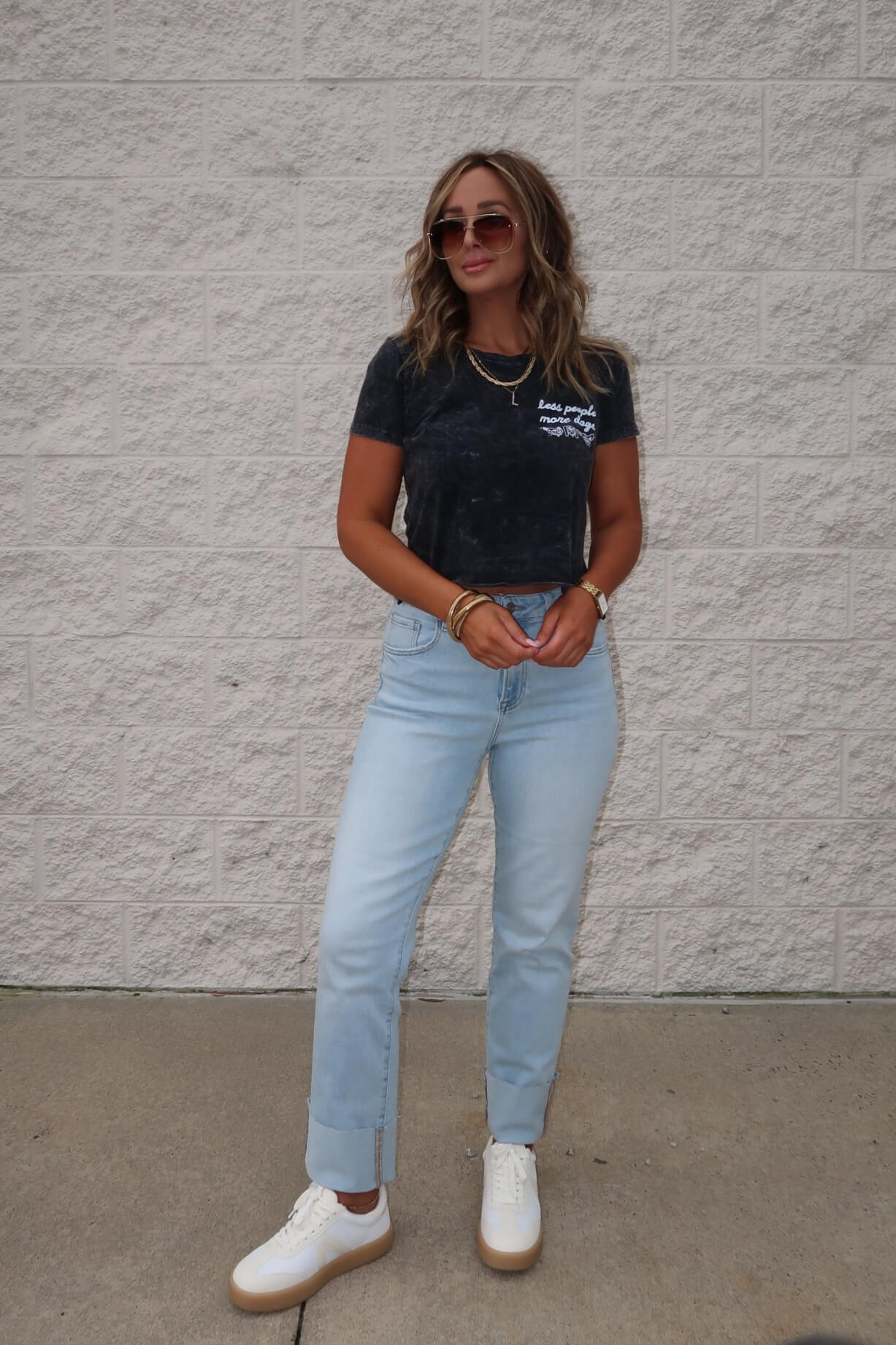 Woman in sunglasses, light jeans, and white sneakers wears the Less People More Dogs Graphic Tee - Black in front of a textured wall.