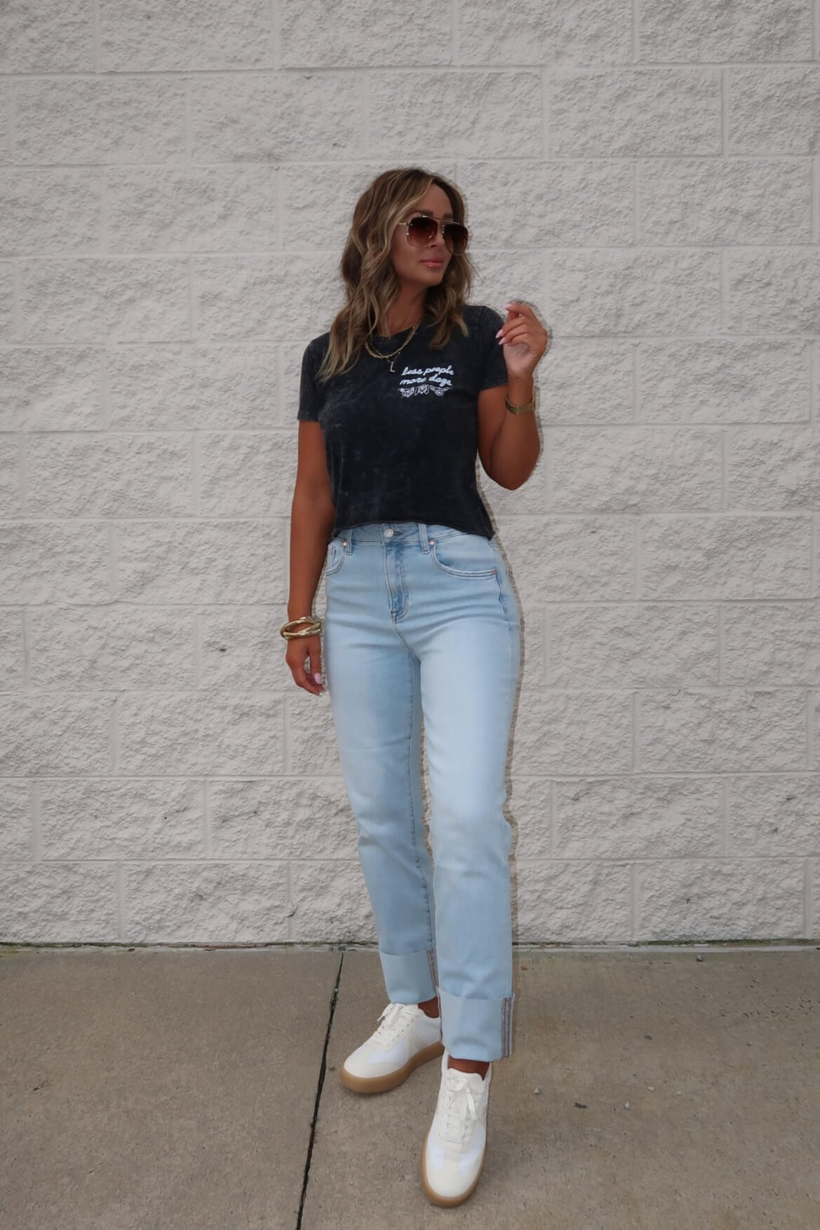 Woman in sunglasses, black Less People More Dogs Graphic Tee, light blue jeans, and white sneakers stands by a brick wall.