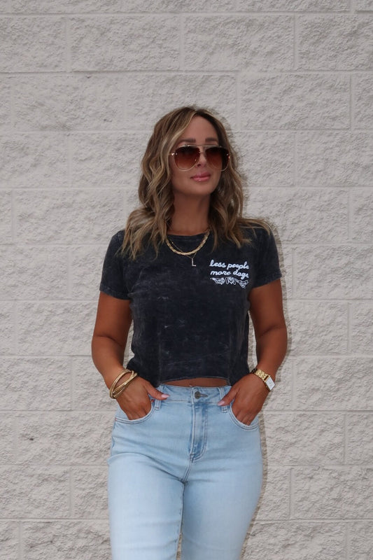 Woman in sunglasses wears the Less People More Dogs Graphic Tee in black and light blue jeans, standing by a light brick wall.