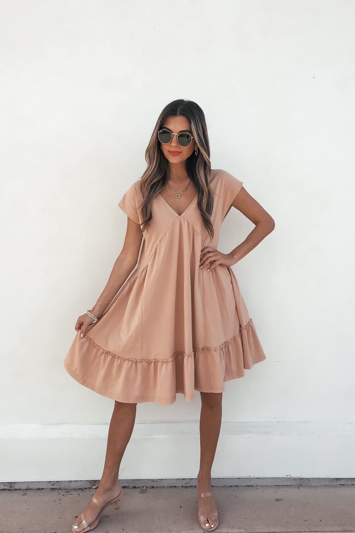 Woman in a Light Apricot V Neck Mini Dress with ruffled sleeves, sunglasses, and heels poses against a plain white background.