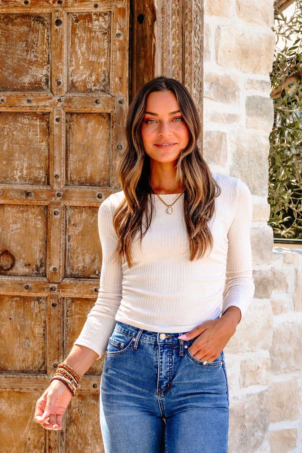 Woman with long brown hair wears a Light Beige Ruched Side Ribbed Top and blue jeans, smiling by a rustic door and stone wall.