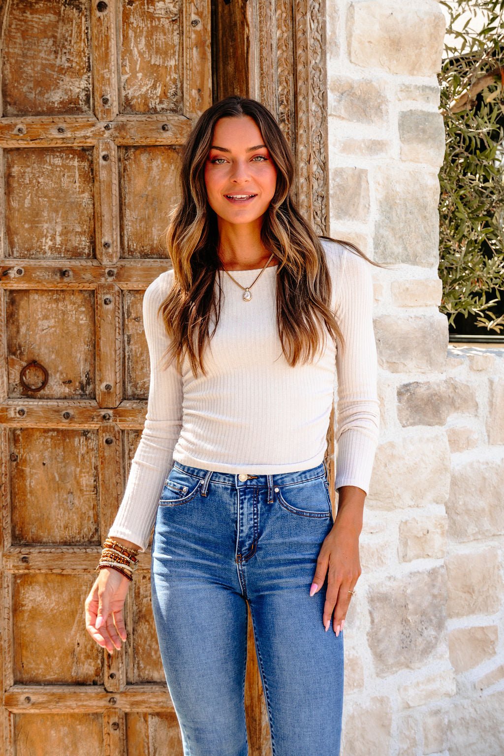Woman with long wavy hair in a Light Beige Ruched Side Ribbed Top and blue jeans smiles by a rustic wooden door and stone wall.