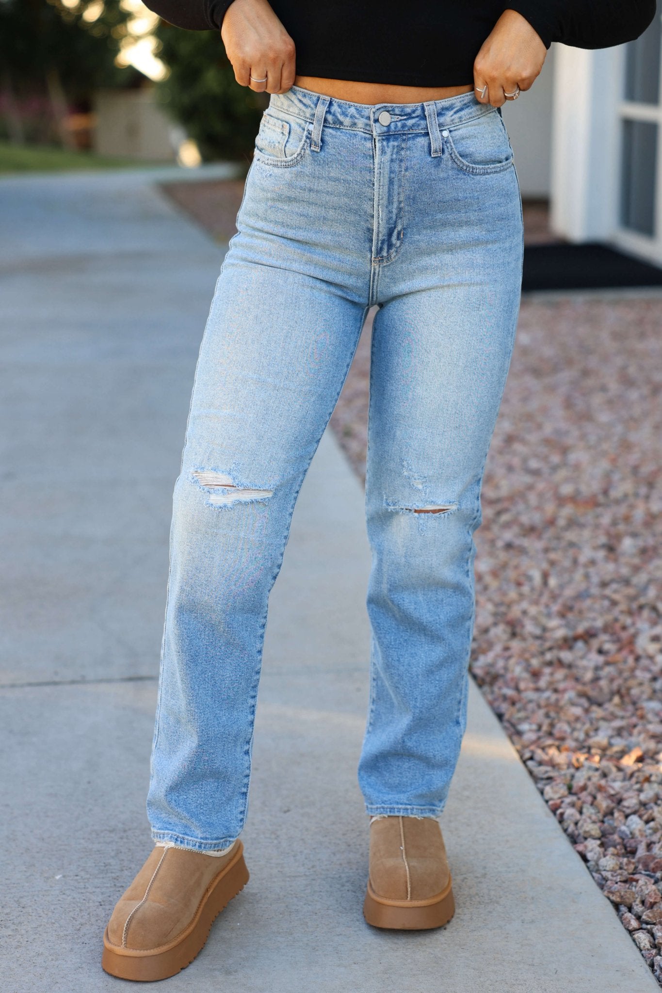 A person in Light Denim Distressed High Rise Dad Jeans, a black top, and tan platform shoes stands on a sidewalk by gravel.