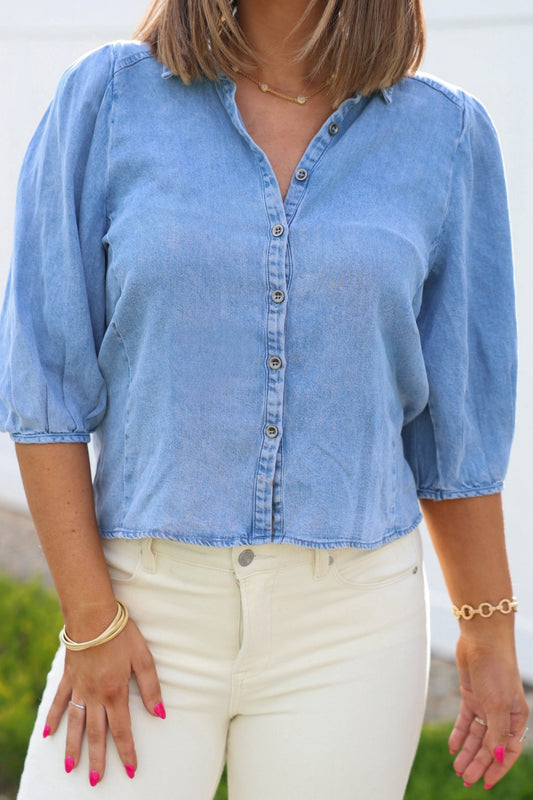 A woman in a Light Denim Pleated Button Down Shirt, high-waisted white pants, gold jewelry, and pink nail polish.