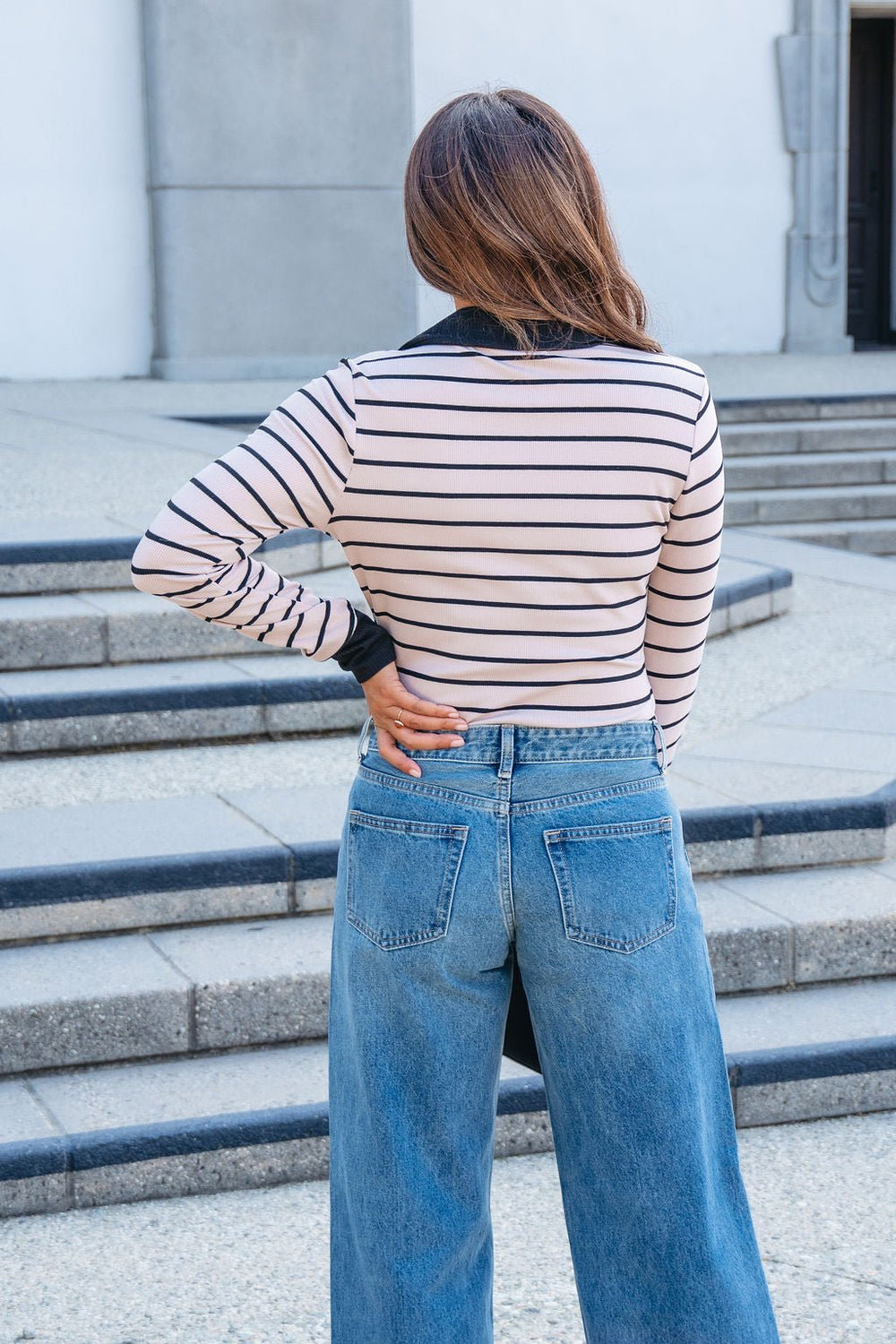 A woman stands outdoors near concrete steps in a Light Mocha Striped Rib Knit Top and blue jeans, facing away.