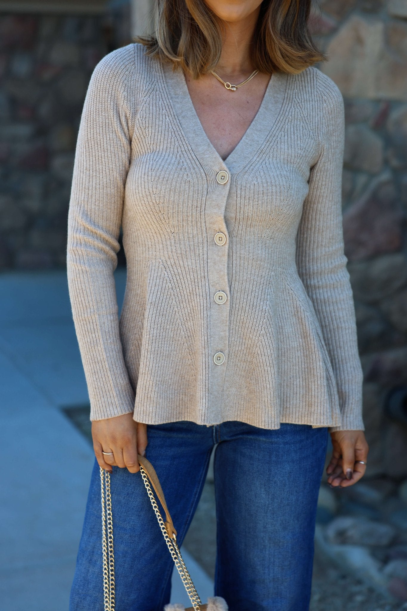 Woman in Light Tan Hourglass Sweater Cardigan with buttons, blue jeans, and a chain strap bag, standing outdoors.