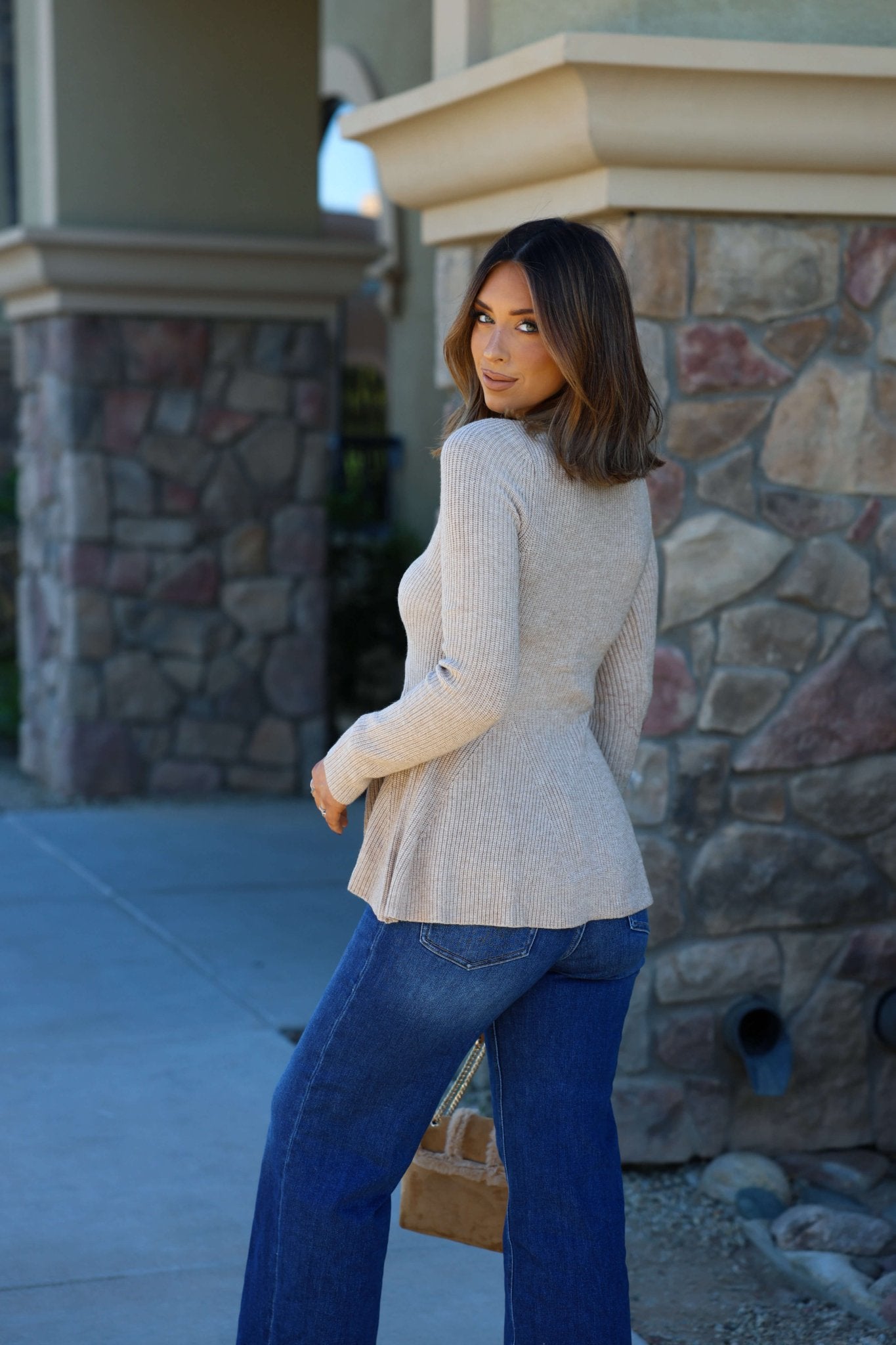 A woman in a Light Tan Hourglass Sweater Cardigan and blue jeans stands outdoors, glancing back near a stone building.