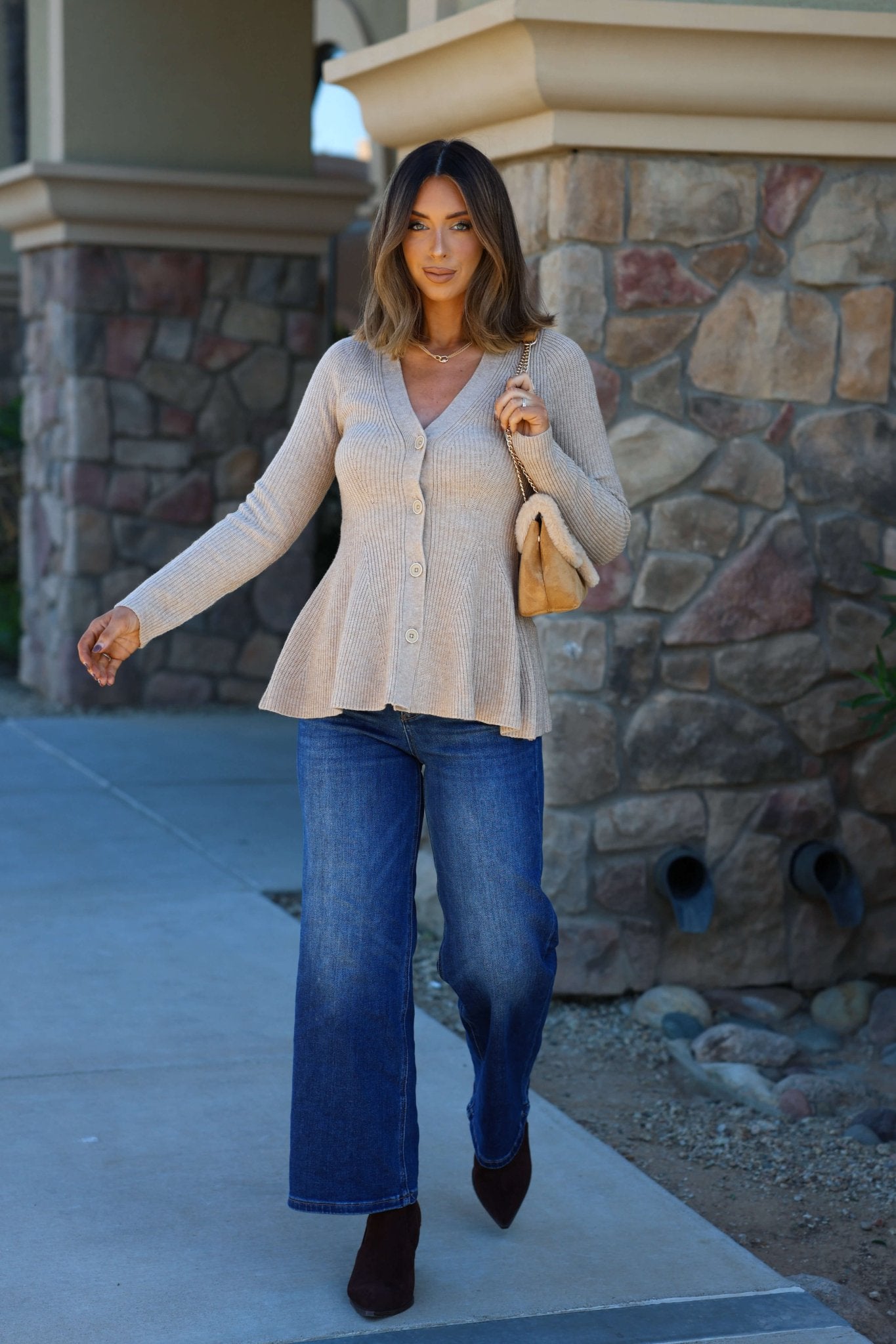 Woman outdoors in a light tan hourglass sweater cardigan, blue wide-leg jeans, brown shoes, and holding a tan purse.