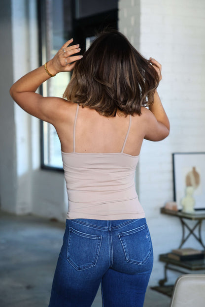 A woman with shoulder-length brown hair wears the Light Taupe Seamless Cami Tank Top and blue jeans indoors, ideal for layering.
