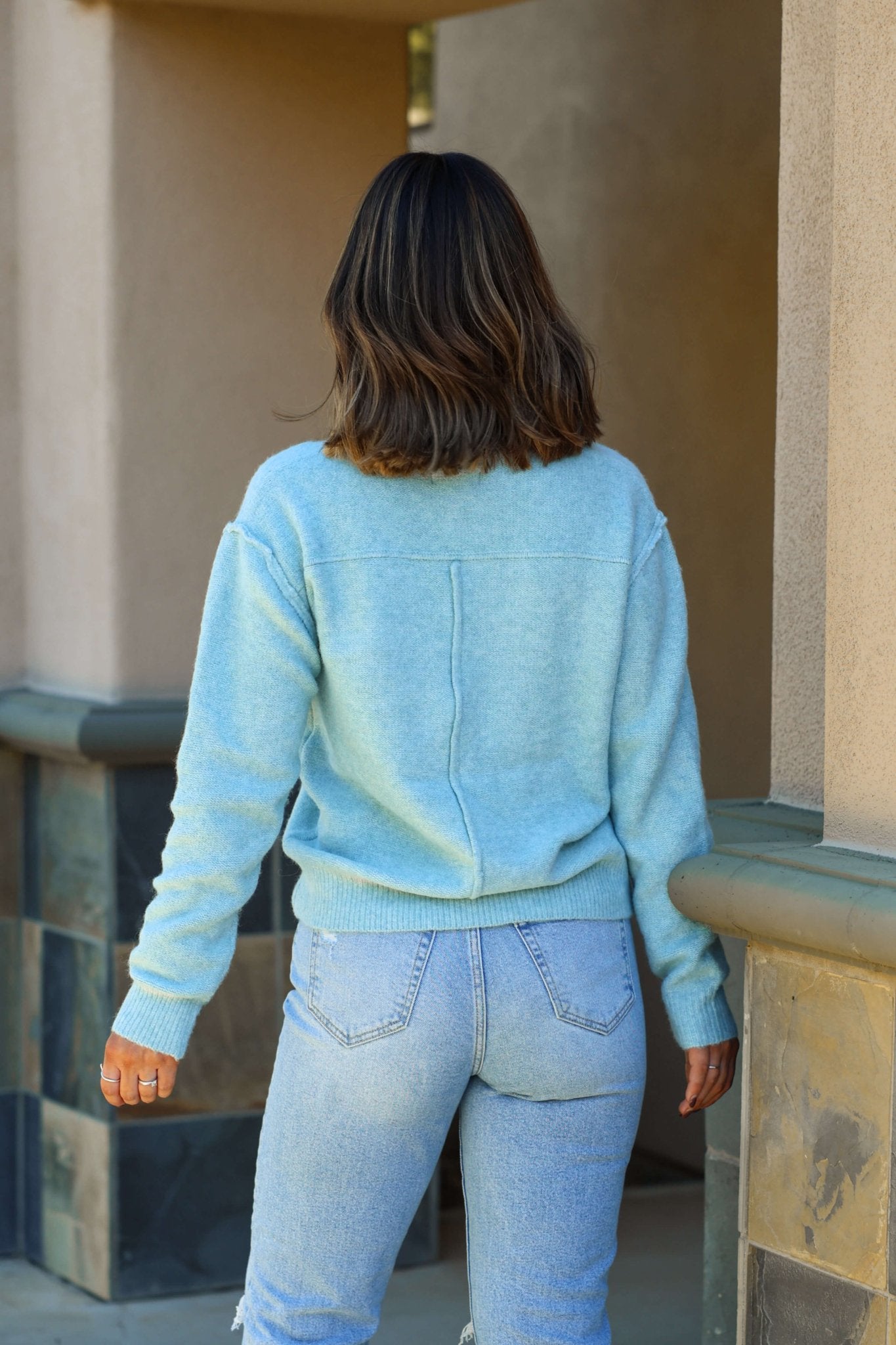 A person with shoulder-length brown hair in the Light Teal Cozy V Neck Sweater stands outdoors facing away.