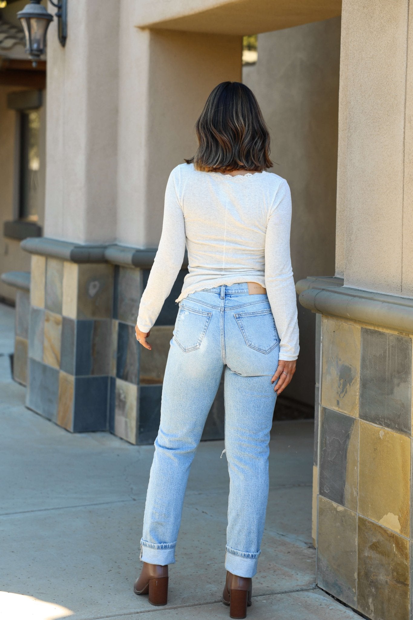 A woman stands outside, facing away, wearing a light top and Light Wash Distressed Straight Cuffed Jeans.