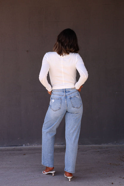 A woman stands with her back to the camera, wearing a white top and Light Wash Mid Rise Distressed Dad Jeans with white heeled sandals.
