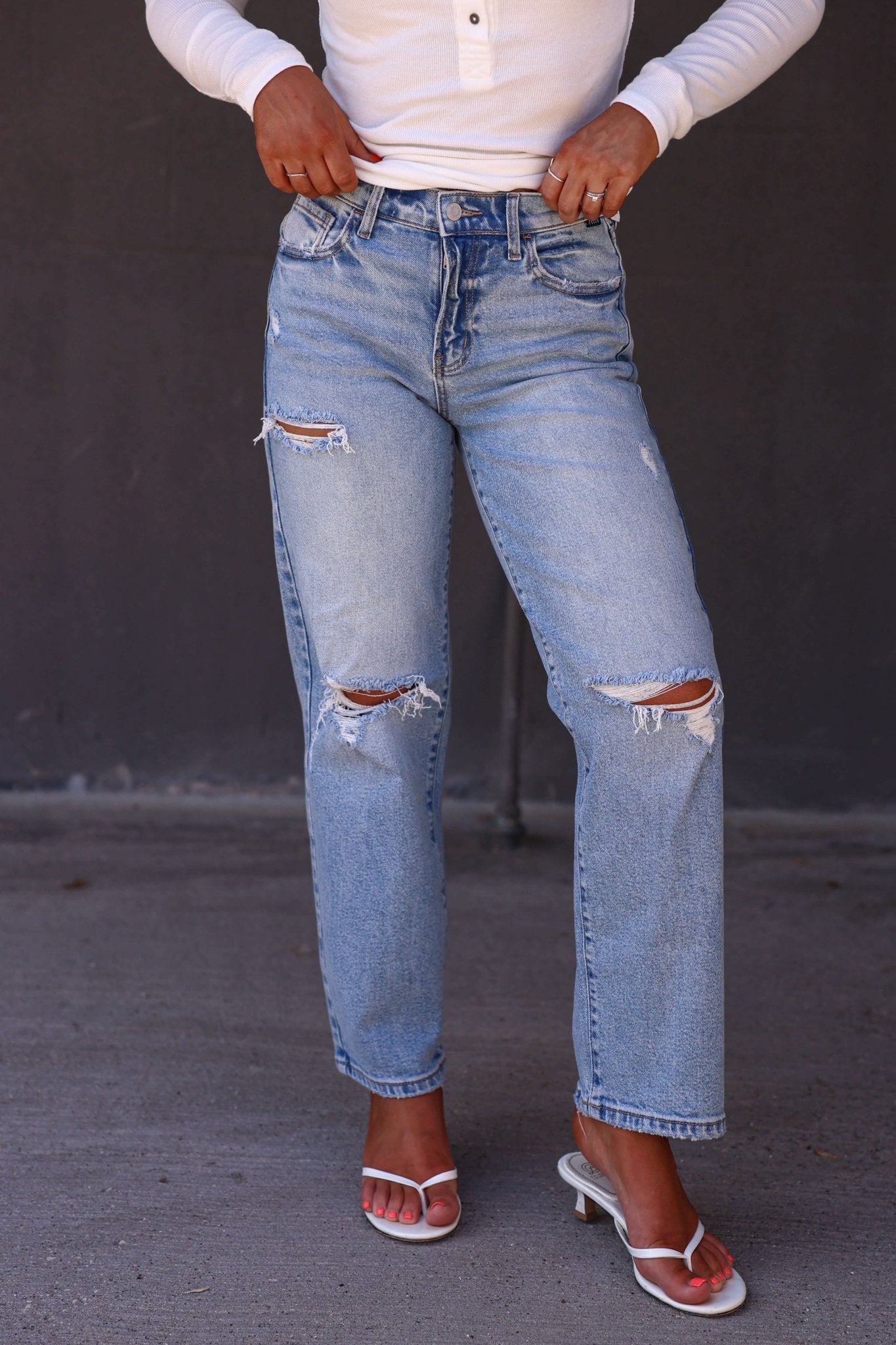 Wearing Light Wash Mid Rise Distressed Dad Jeans, a white long-sleeve top, and white heeled sandals on a gray surface.