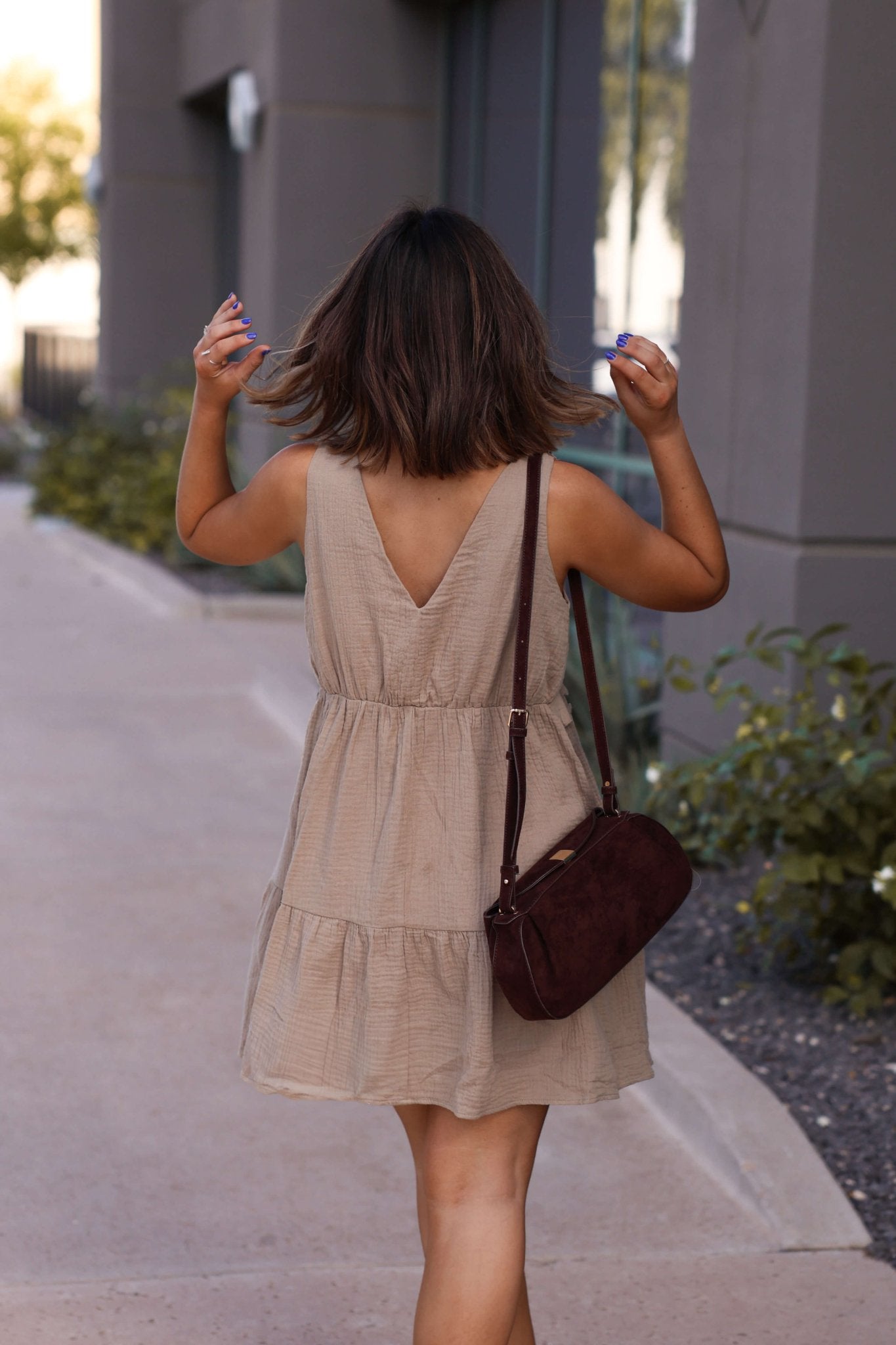A woman in the Lila Mocha Ruffle Tiered Tank Mini Dress walks on a sidewalk, her back to the camera, carrying a dark brown bag.