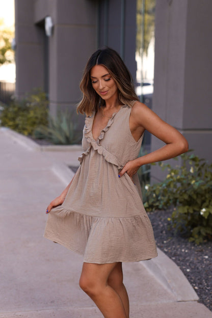 A woman stands on the sidewalk in the Lila Mocha Ruffle Tiered Tank Mini Dress - DOORBUSTER, looking down and smiling.
