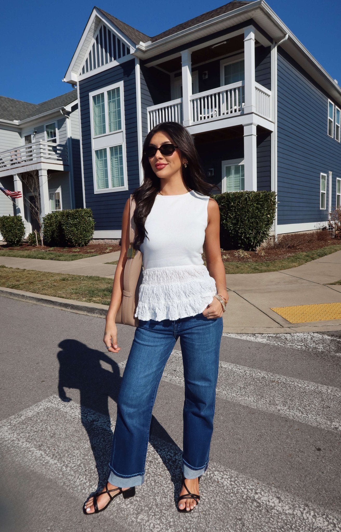 Woman in sunglasses wearing the Lilly White Lace Peplum Top and blue jeans stands on a street with blue and white houses.