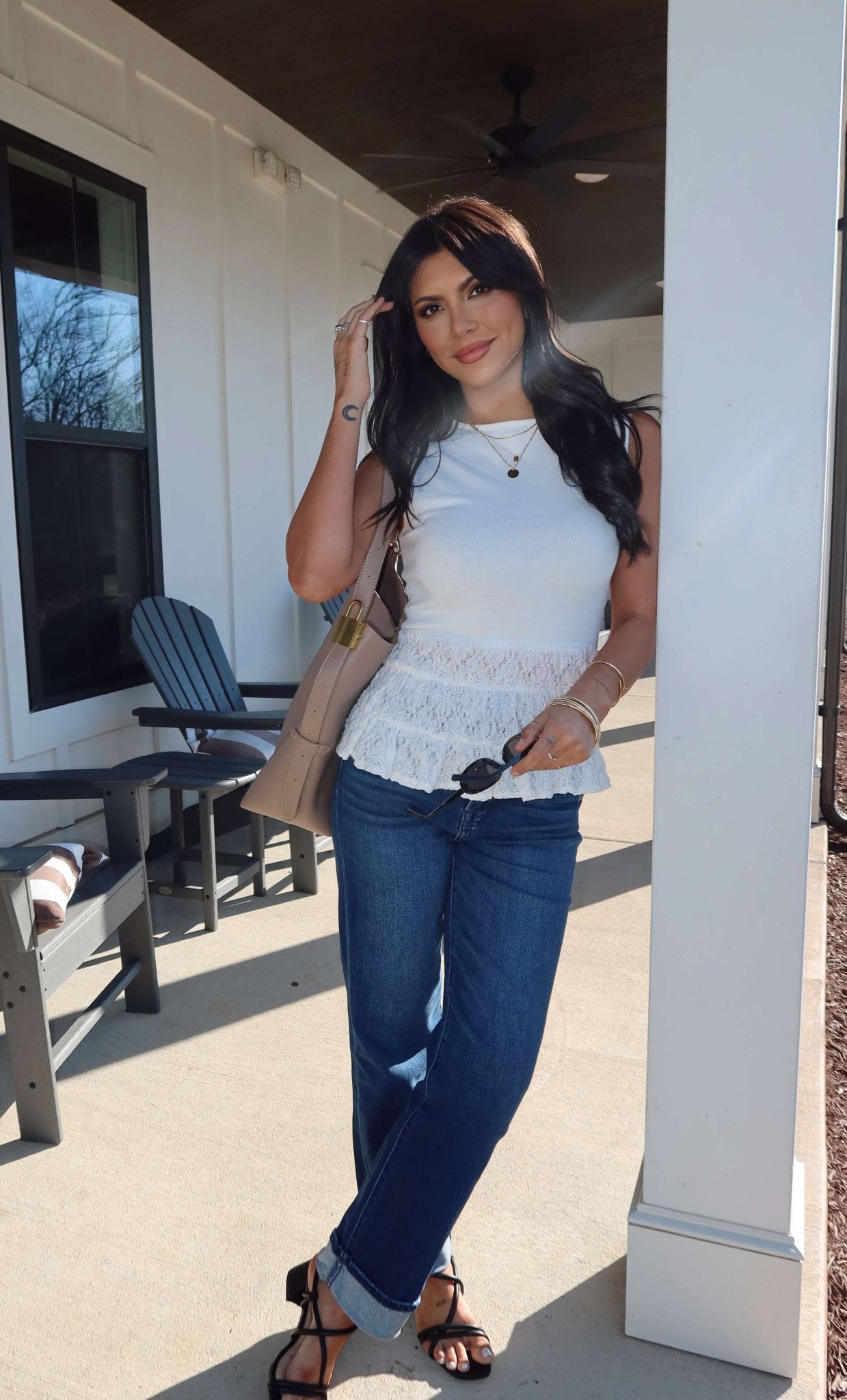 Woman wears the Lilly White Lace Peplum Top with jeans, holding sunglasses and a purse while leaning on a porch column.
