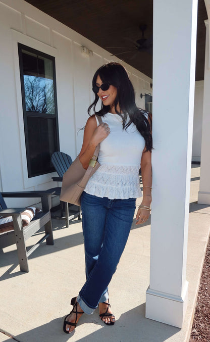 Woman in sunglasses outdoors leans on a pillar, wearing the Lilly White Lace Peplum Top with jeans and sandals, holding a beige bag.