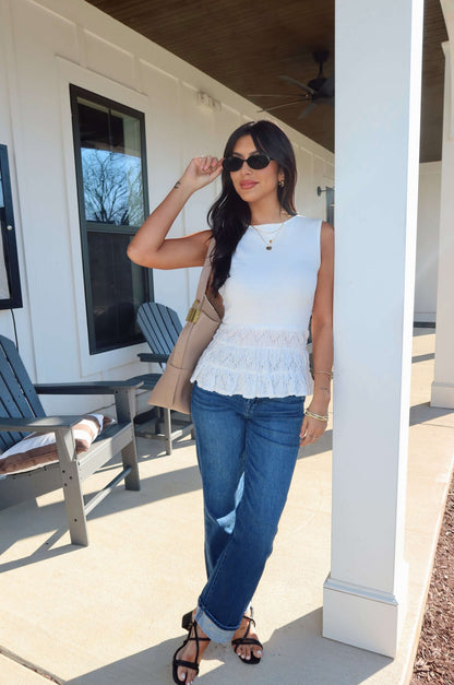Woman wearing the Lilly White Lace Peplum Top, sunglasses, and blue jeans, holding a beige handbag by a white pillar outdoors.
