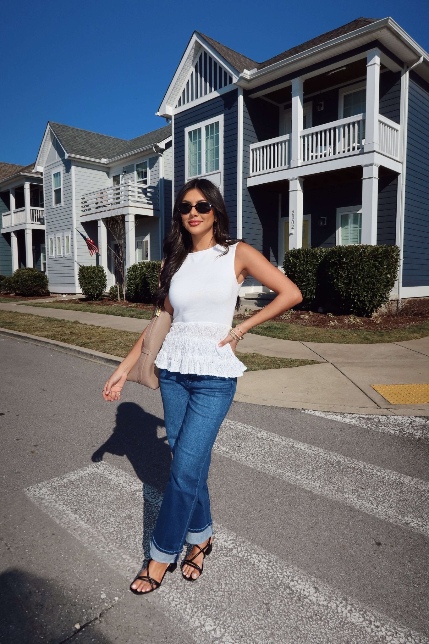 A woman in a Lilly White Lace Peplum Top and jeans stands on a crosswalk before modern blue houses on a sunny day.