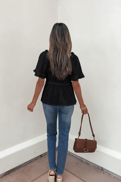Woman with long hair wears the Lola Black Ruffled Babydoll Top and blue jeans, holding a brown purse while facing a white wall.