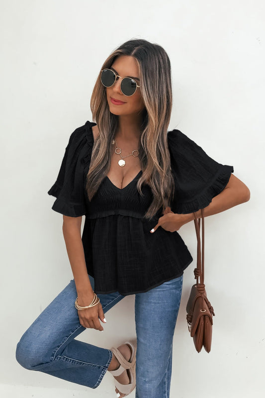 Woman in sunglasses wears the Lola Black Ruffled Babydoll Top with jeans and holds a brown purse against a white wall.