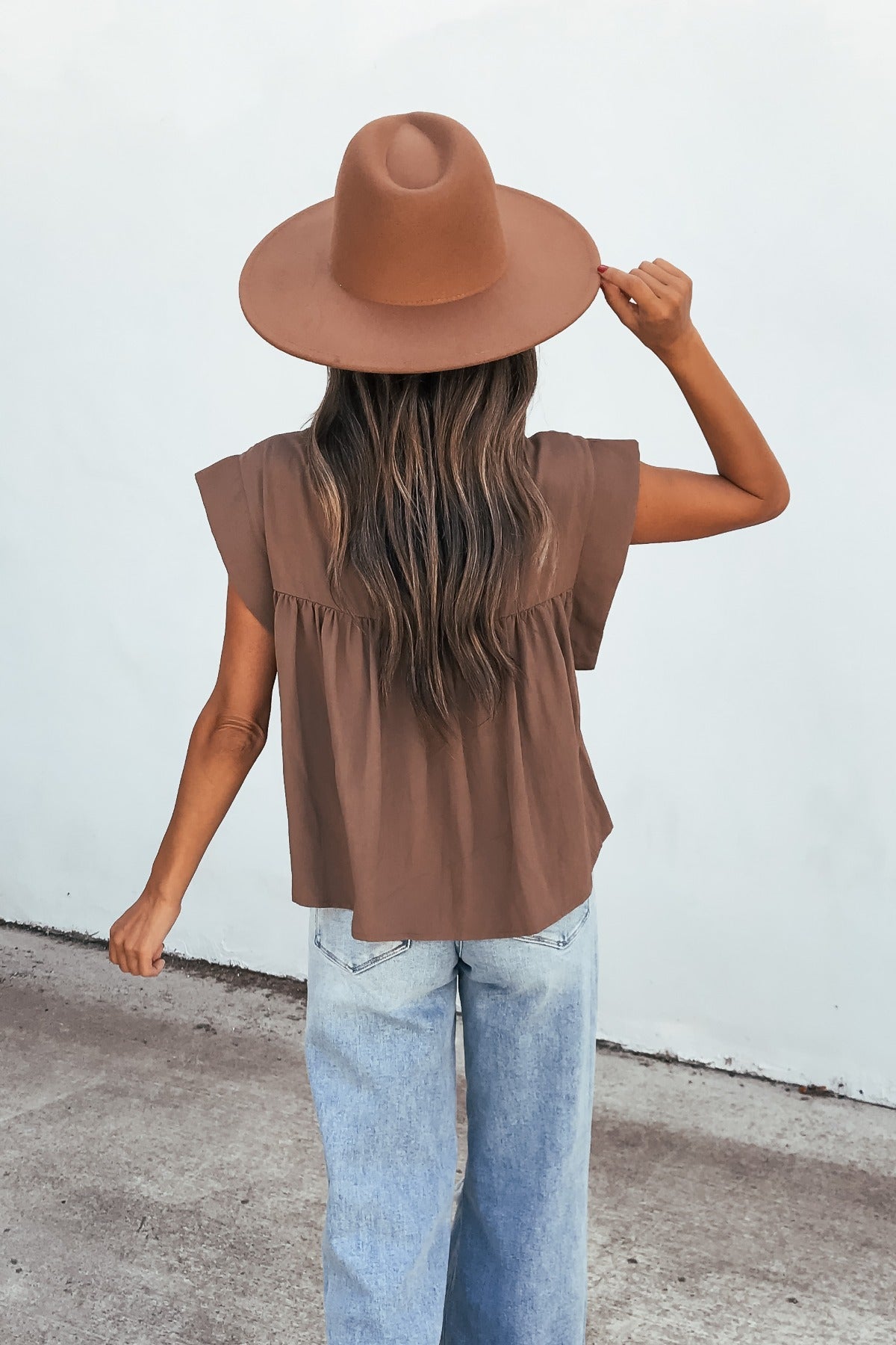Woman with long hair in a wide-brimmed brown hat, Lola Button Up Lace Top - Mocha, and light blue jeans stands with back to camera.
