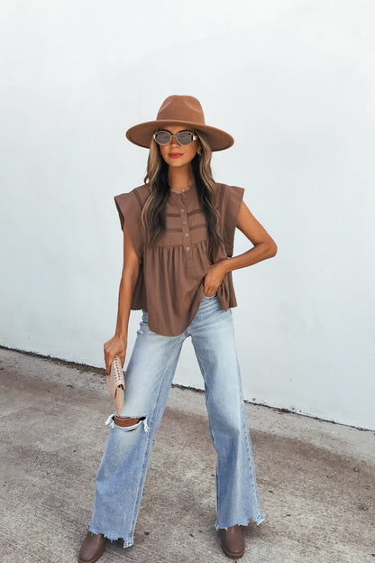 Woman in a brown hat, sunglasses, Lola Button Up Lace Top - Mocha, ripped light jeans, brown shoes, and beige clutch stands by a white wall.