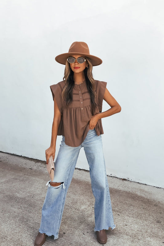 Woman in a brown hat, sunglasses, Lola Button Up Lace Top - Mocha, ripped light jeans, brown shoes, and beige clutch stands by a white wall.