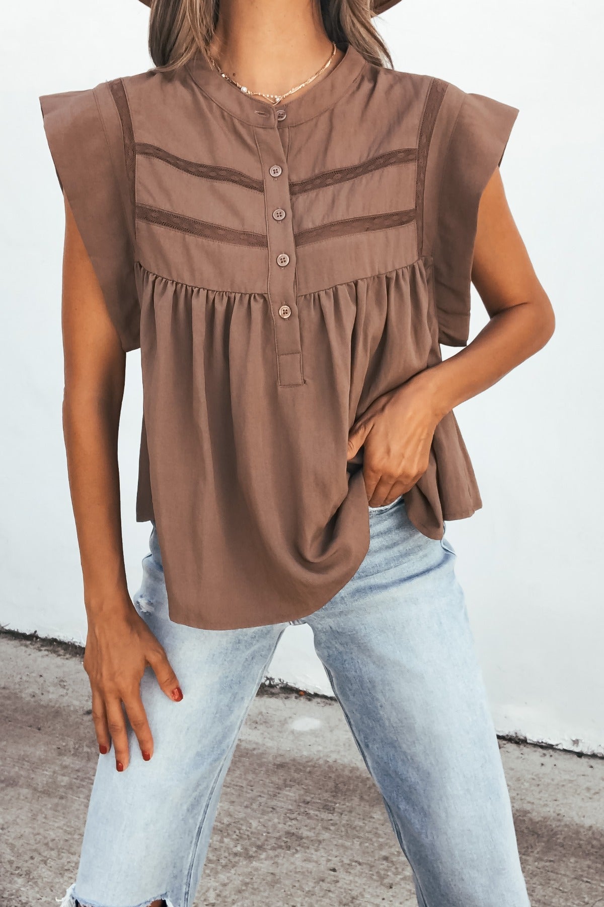 Woman in a Lola Button Up Lace Top - Mocha with light blue jeans stands outdoors against a white wall.