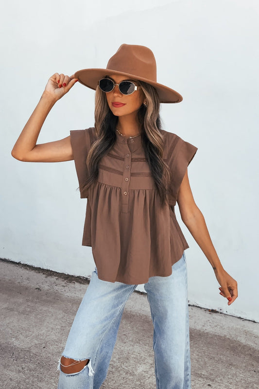 Woman in a brown hat, sunglasses, Lola Button Up Lace Top - Mocha, and ripped jeans poses by a white outdoor wall.