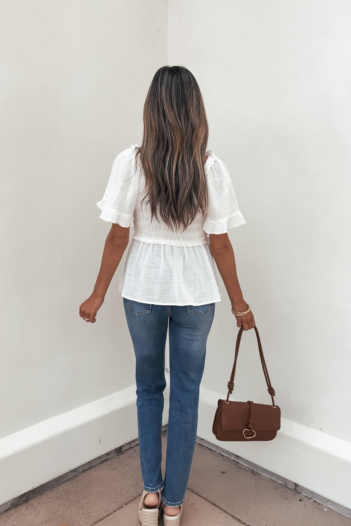Woman with long hair wears the Lola White Ruffled Babydoll Top and blue jeans, holding a brown handbag while facing a white wall.