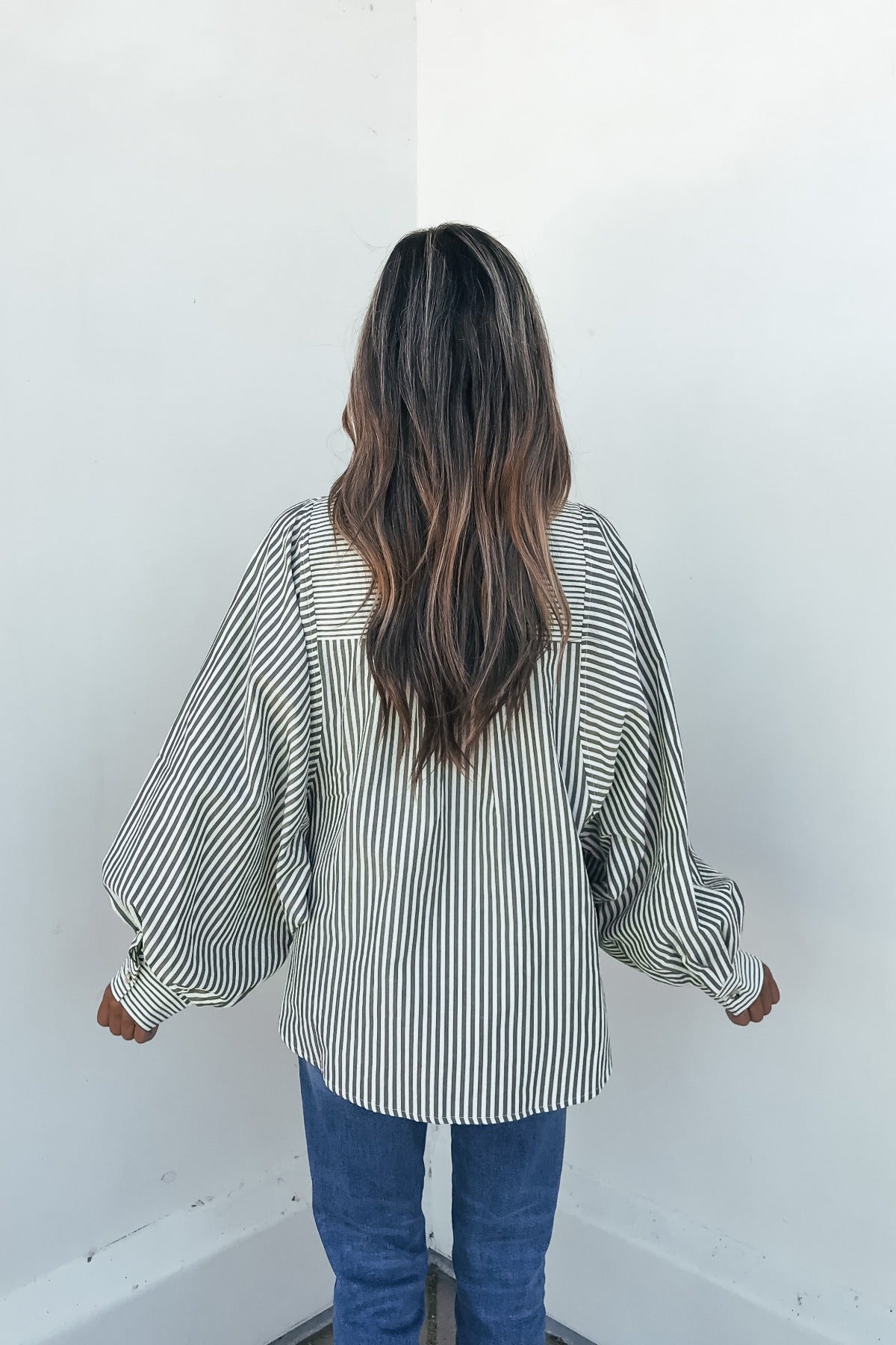 Woman in the Loose Ends Striped Shirt - Olive and blue jeans stands facing a white corner wall.