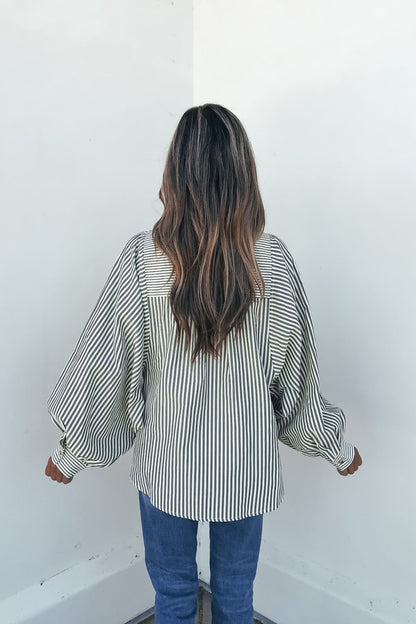 Woman in the Loose Ends Striped Shirt - Olive and blue jeans stands facing a white corner wall.