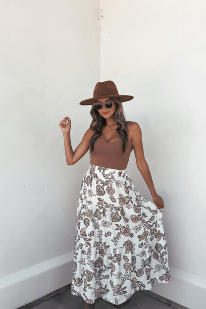 Woman in a brown hat, sunglasses, brown top, and Lorelei Brown Floral Print Midi Skirt stands against a white wall holding her skirt.