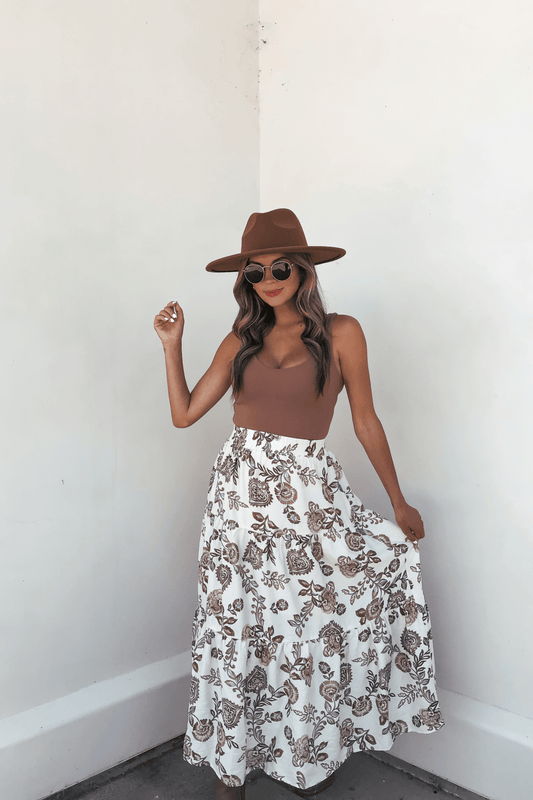 Woman in a brown hat, sunglasses, brown top, and Lorelei Brown Floral Print Midi Skirt stands against a white wall holding her skirt.