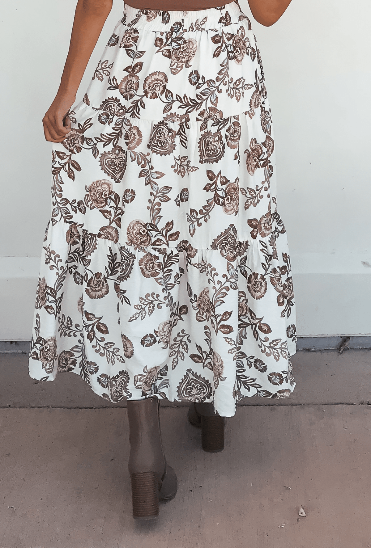 A person wearing the Lorelei Brown Floral Print Midi Skirt with brown heeled boots, standing on a concrete surface.