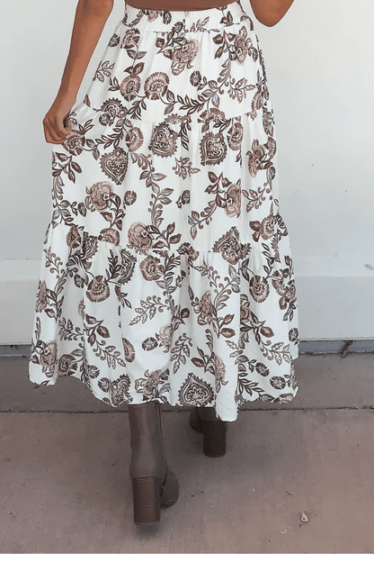 A person wearing the Lorelei Brown Floral Print Midi Skirt with brown heeled boots, standing on a concrete surface.