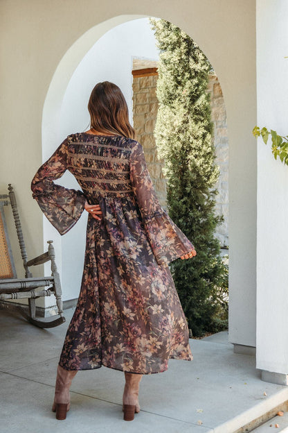 Woman in the Lori Brown Floral Print Smocked Midi Dress and heeled boots stands outdoors under an archway, back to the camera.