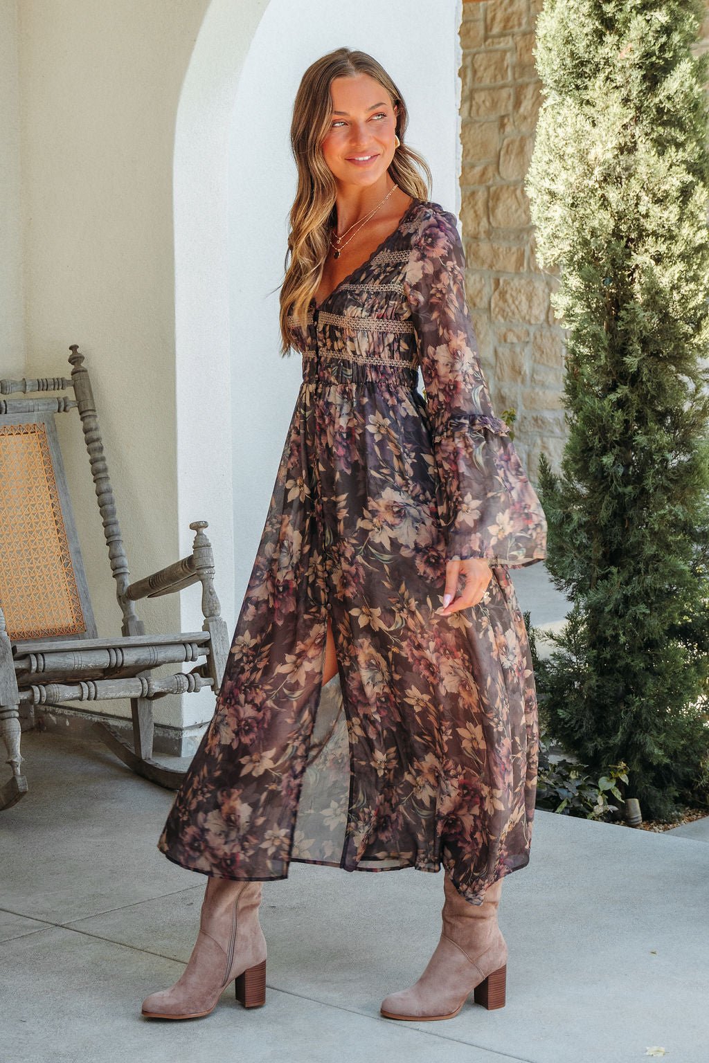 Woman in Lori Brown Floral Print Smocked Midi Dress smiles outdoors by a wooden chair and lush green bush.