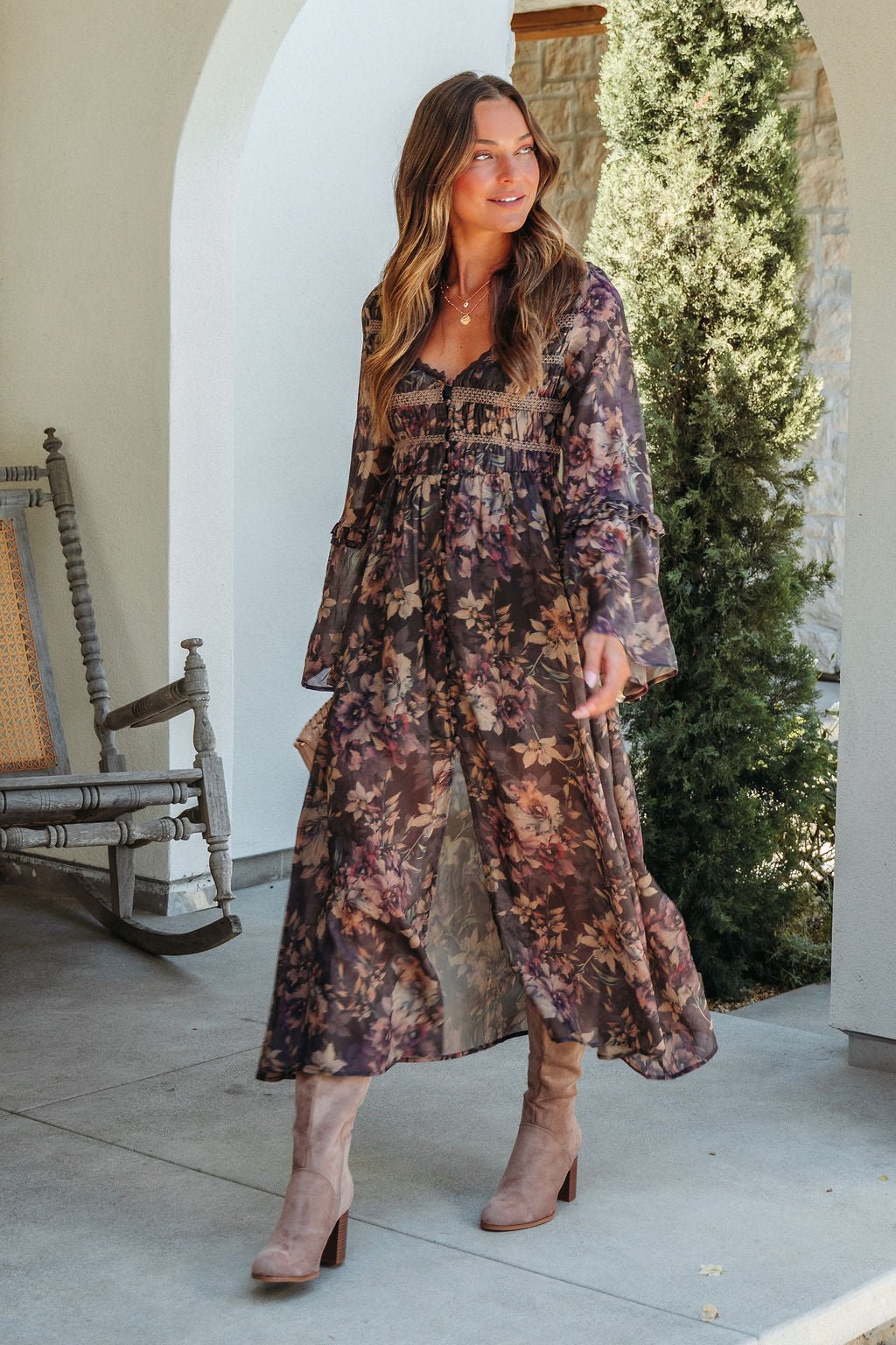 Woman in the Lori Brown Floral Print Smocked Midi Dress walks outdoors by a rocking chair and lush greenery.