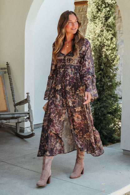 Woman in the Lori Brown Floral Print Smocked Midi Dress walks outdoors by a rocking chair and lush greenery.