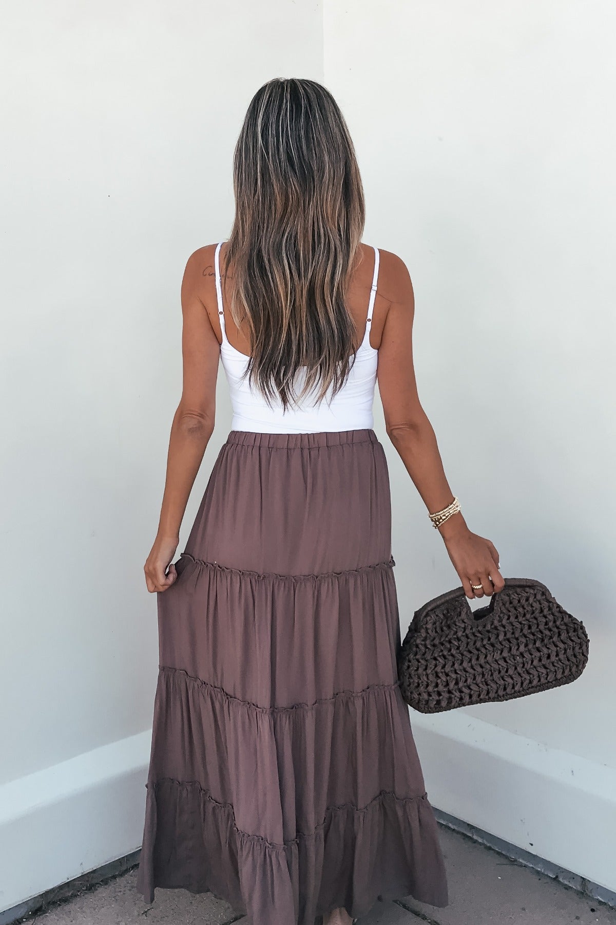Woman with long hair in a white tank top and Lori Ruffle Tiered Maxi Skirt - Mocha, holding a textured brown handbag by a white wall.