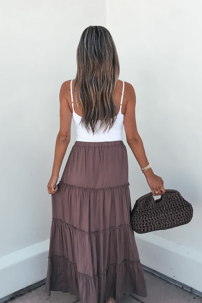 Woman with long hair in a white tank top and Lori Ruffle Tiered Maxi Skirt - Mocha, holding a textured brown handbag by a white wall.