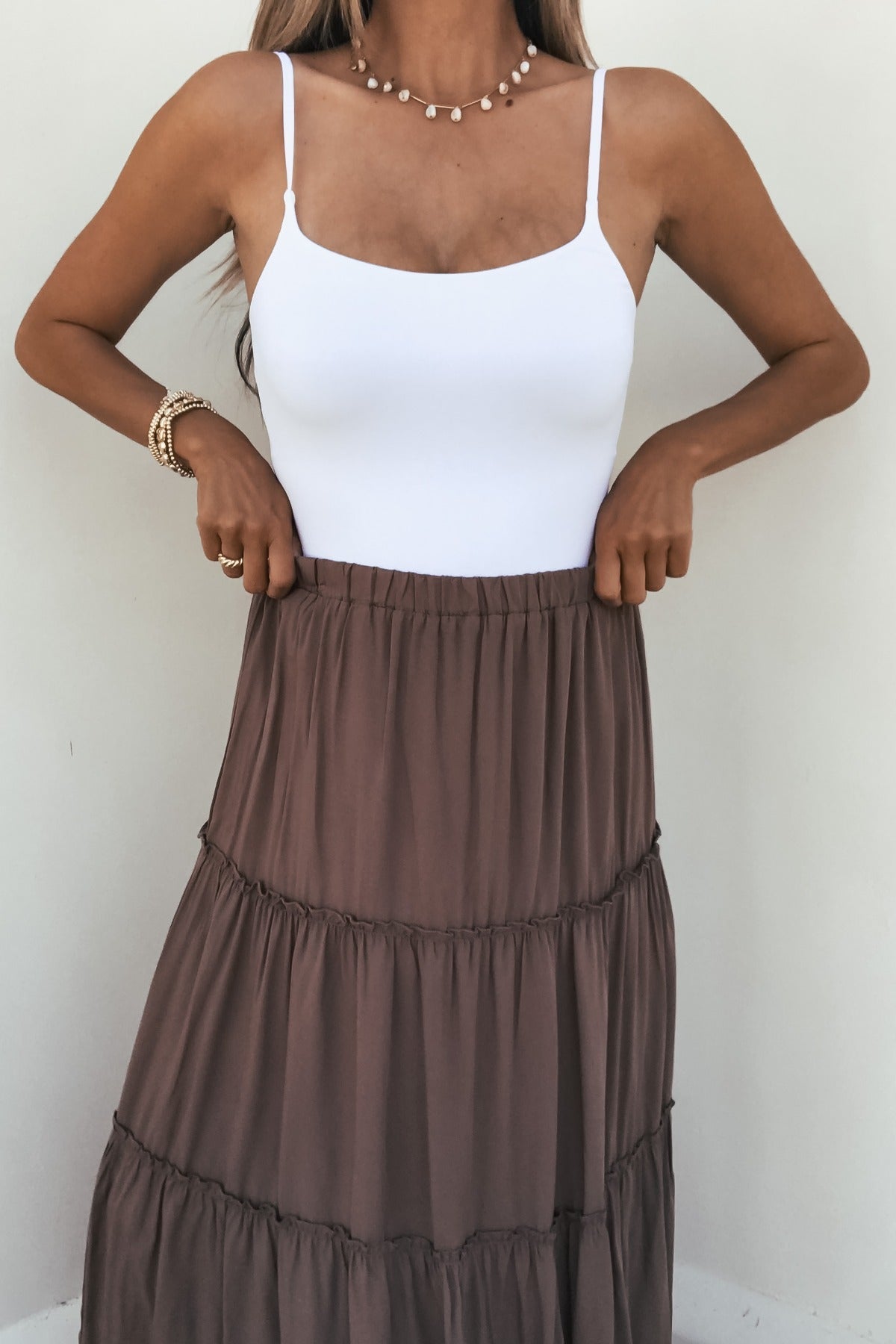 Woman in a white tank top, pearl necklace, and Lori Ruffle Tiered Maxi Skirt - Mocha, standing against a plain background.